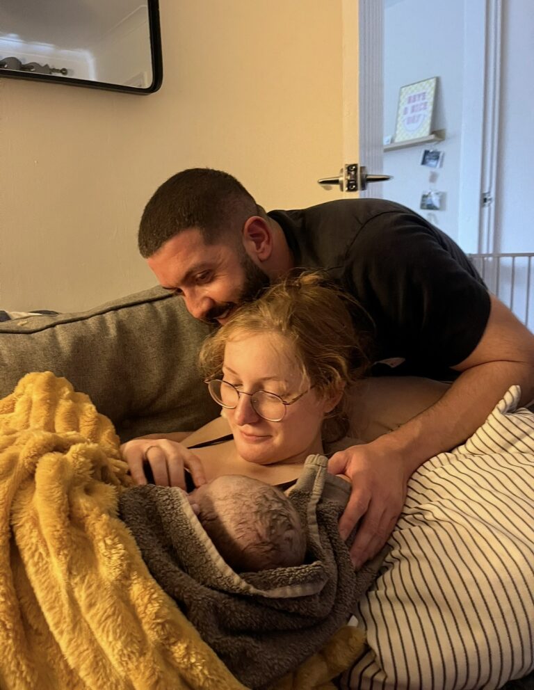 A new mum on the sofa holds her baby wrapped in towels whilst her male partner cradles her shoulder and looks at the baby